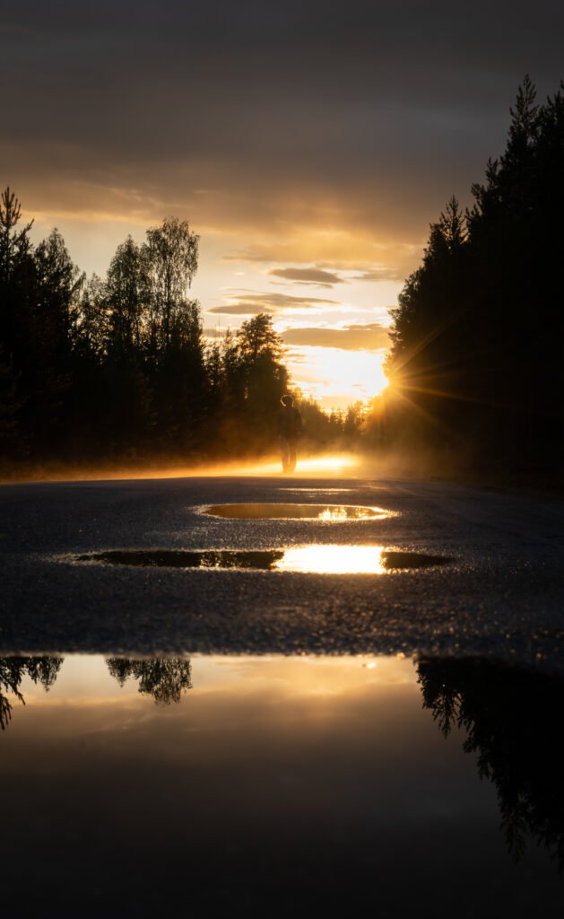 Vrouw loopt over de weg met waterplassen, de ondergaande zon op de achtergrond weerspiegeld in de waterplassen tijdens InnerTrail Sweden